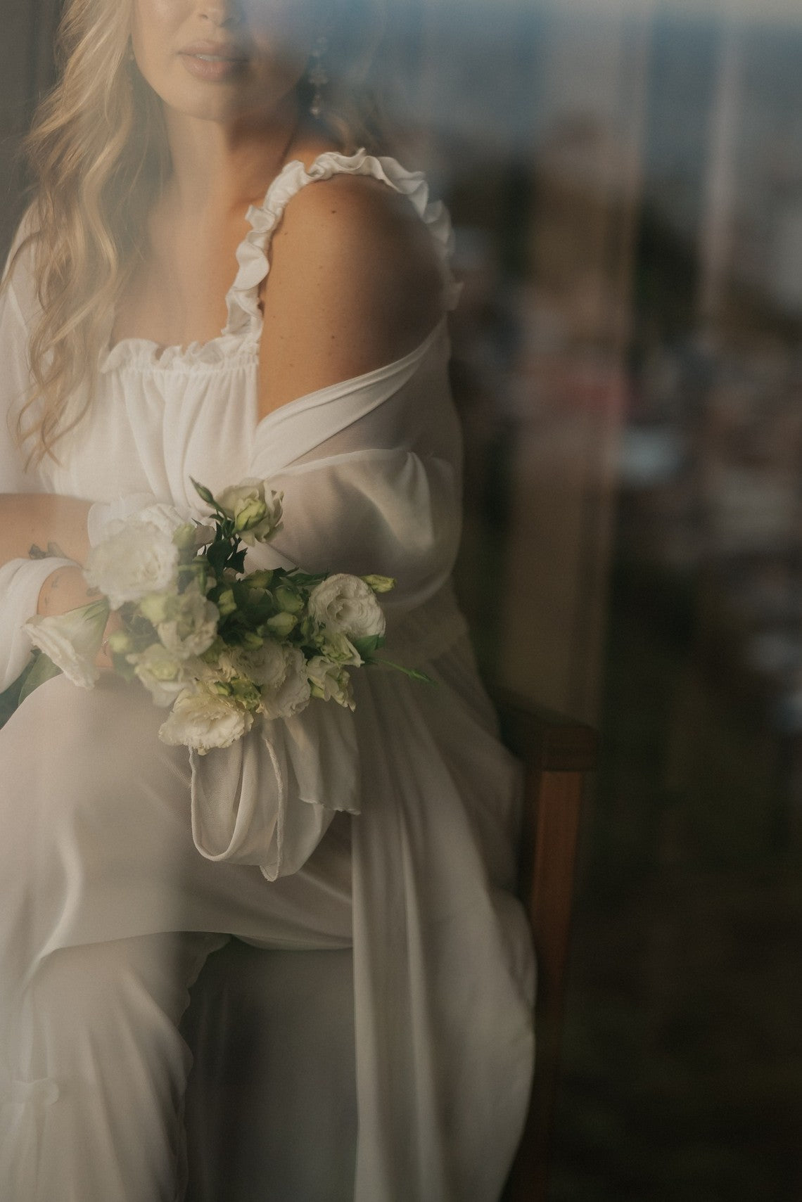 Joana Costa vista através do vidro, com robe de tule e um buquê de flores brancas
